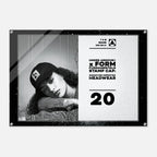 A rectangular black-framed acrylic wall sign featuring a black-and-white portrait of a woman wearing a cap on the left and printed text on the right, including 'UNDER ARMOUR' and 'FORM FUNDAMENTALS STAMP CAP SIGNATURE IMPERFECT HEADWEAR' with the number 20, shown against a light gray background.