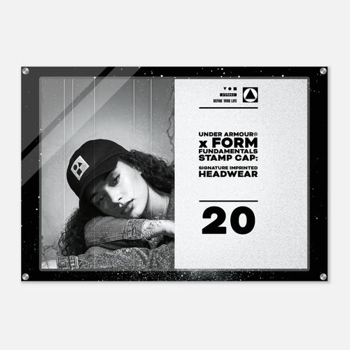 A rectangular black-framed acrylic wall sign featuring a black-and-white portrait of a woman wearing a cap on the left and printed text on the right, including 'UNDER ARMOUR' and 'FORM FUNDAMENTALS STAMP CAP SIGNATURE IMPERFECT HEADWEAR' with the number 20, shown against a light gray background.