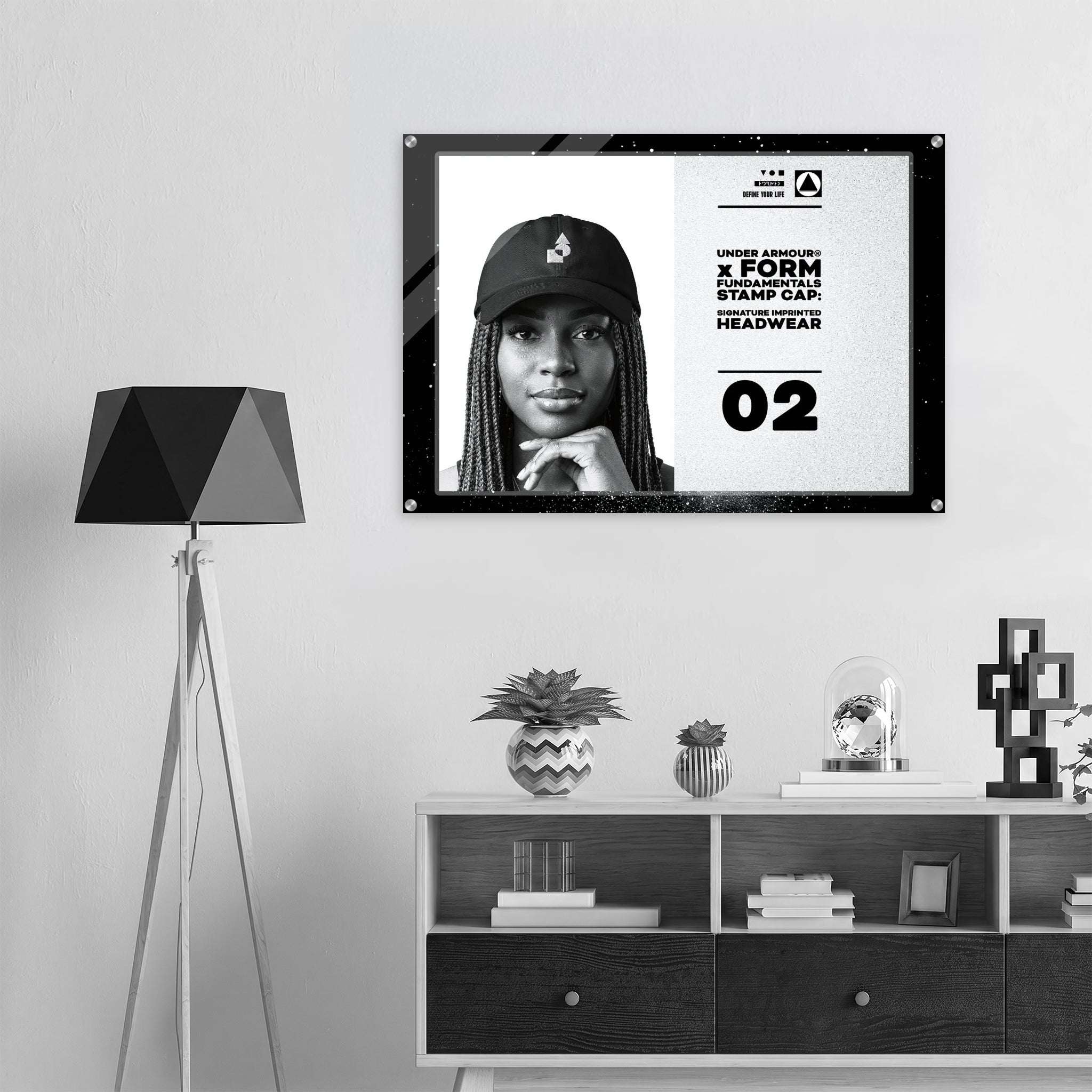 A framed black-and-white poster featuring a portrait of a woman in a cap and bold text, displayed on a light wall above a shelf with decor items. A black tripod floor lamp stands to the left.