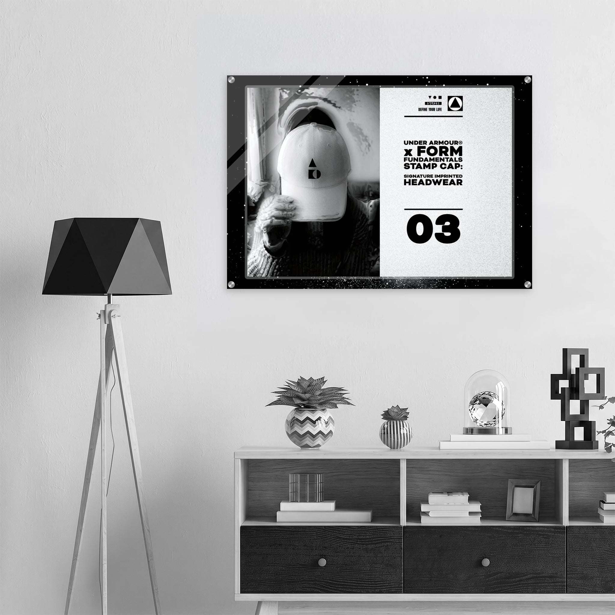 A framed black-and-white poster hanging on a light gray wall above a white and dark wood shelf, with a black tripod floor lamp on the left. The poster features a mirror selfie of a person wearing a white cap and bold text from Form Finding Design.