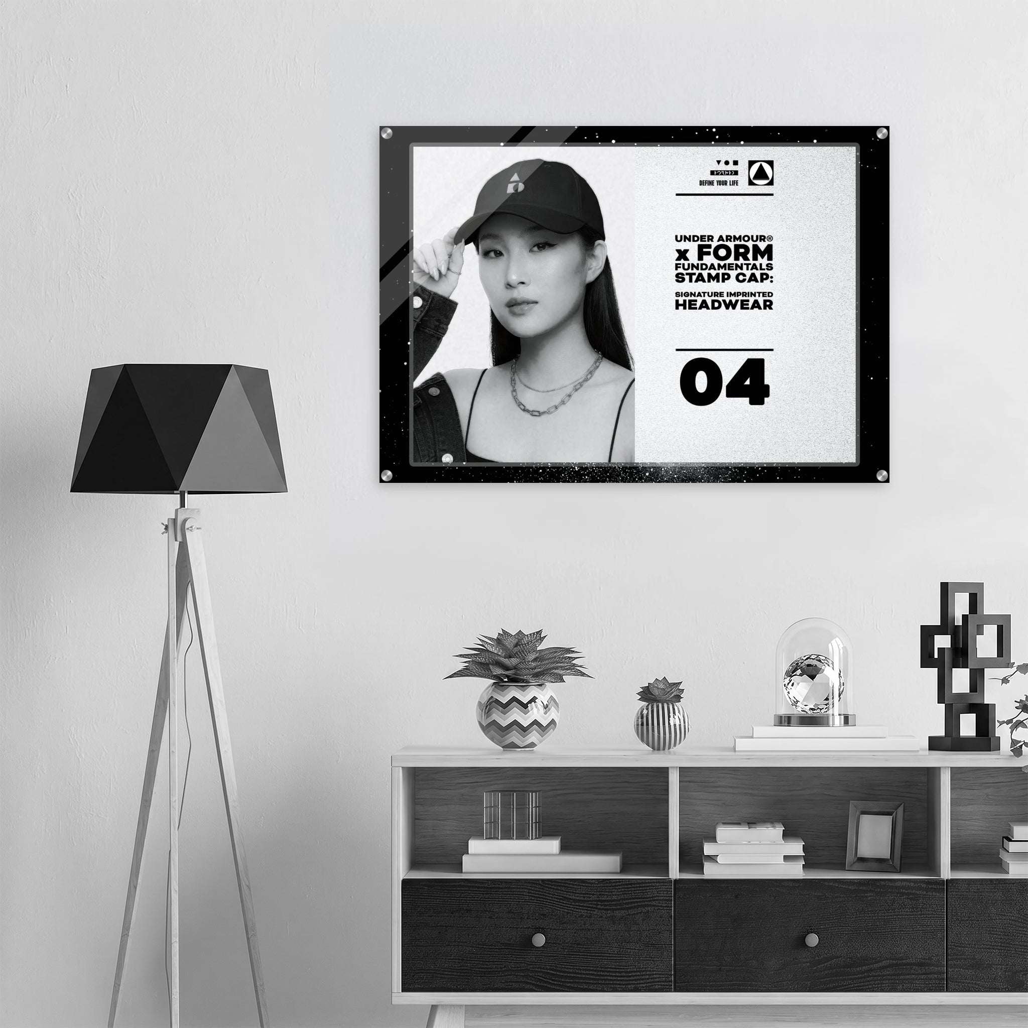 A framed black-and-white promotional print hangs on a light gray wall above a white and dark wood cabinet, next to a black tripod floor lamp. The print shows a woman wearing a baseball cap and includes text from Form Finding Design (FormFD).