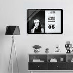 A framed black-and-white poster hangs on a light gray wall above a white shelf with decor items. The poster features a split design: a black photo of a woman wearing a cap on the left, and a white text panel with words from Form Finding Design.