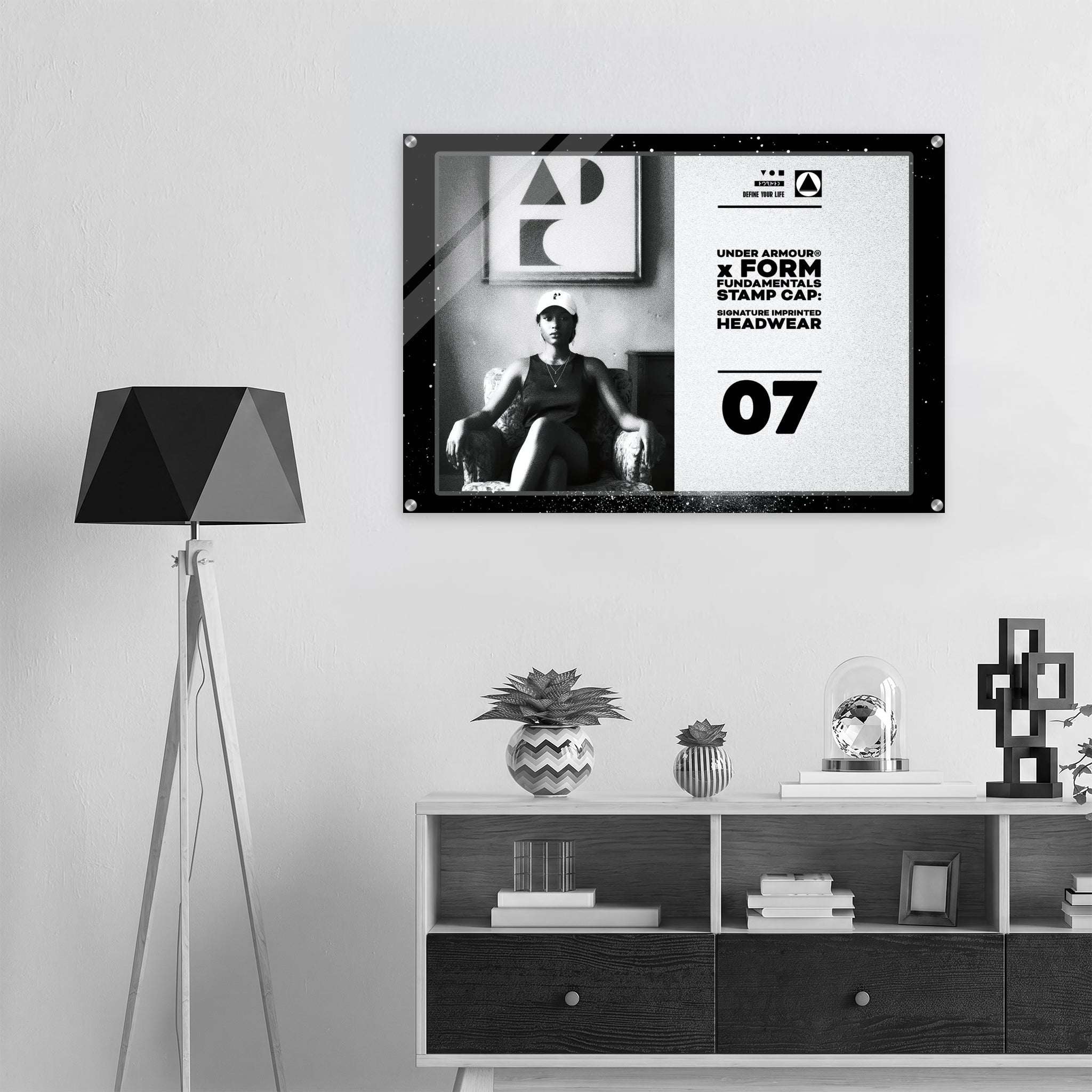 A framed black-and-white poster hangs on a light gray wall above a white and dark wood console shelf, next to a black geometric floor lamp. The poster features a seated person wearing a face mask and text from Form Finding Design (FormFD).