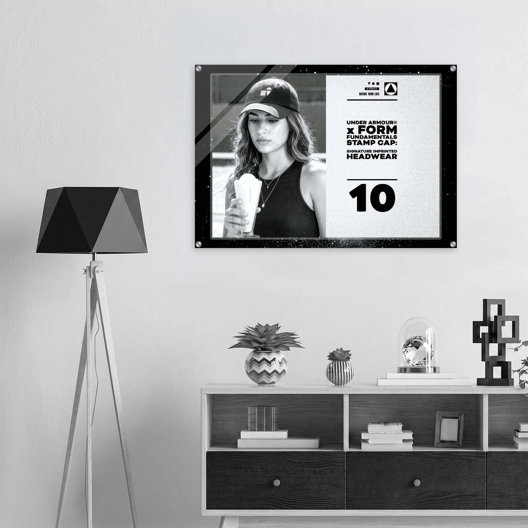 A framed black-and-white poster hanging on a light gray wall above a white and dark wood console shelf, with a black tripod floor lamp on the left. The poster features a black-and-white portrait of a woman in a cap and bold text from Form Finding Design.