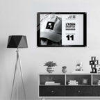 A framed black-and-white poster featuring a person in a cap with bold text and the number 11, displayed on a light gray wall above a white shelf with decor items. A black tripod floor lamp stands to the left.