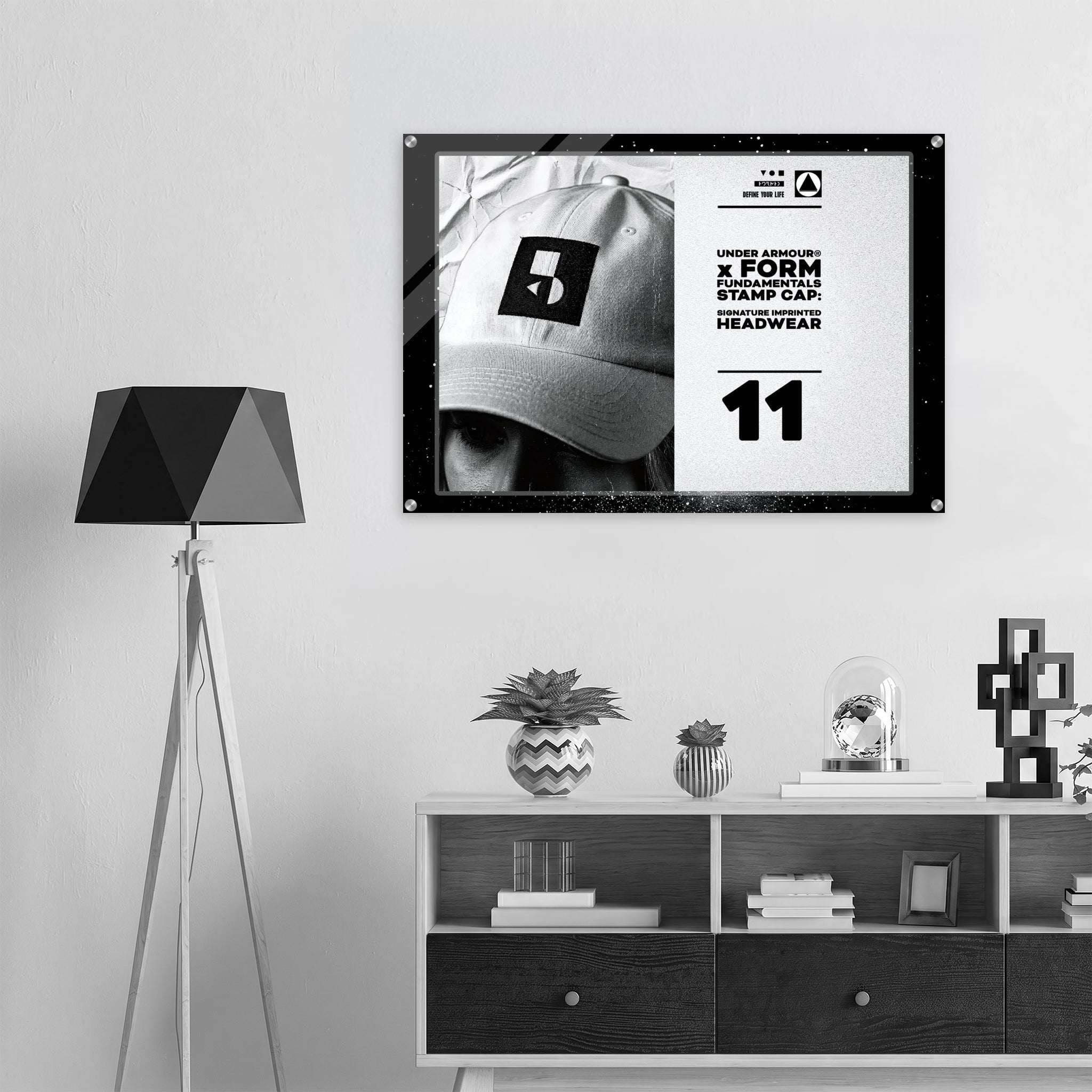 A framed black-and-white poster featuring a person in a cap with bold text and the number 11, displayed on a light gray wall above a white shelf with decor items. A black tripod floor lamp stands to the left.