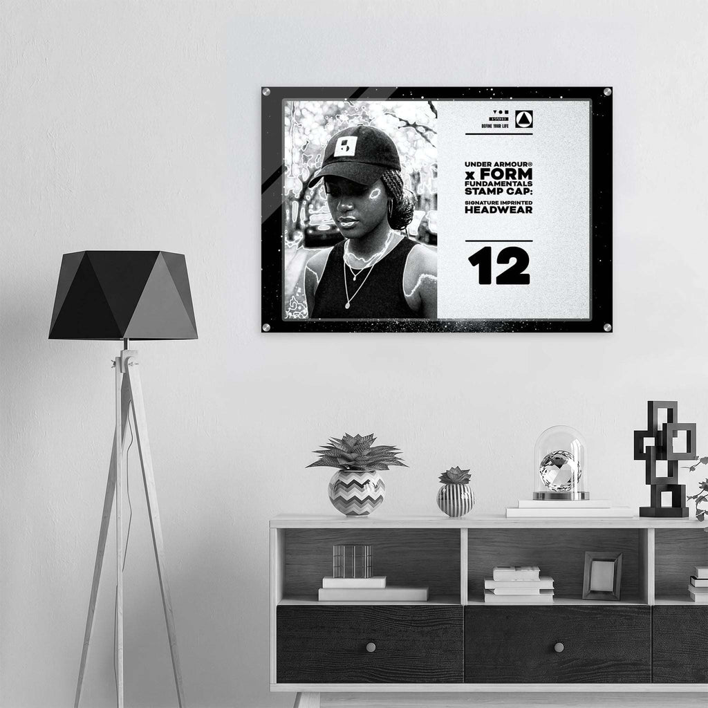A framed black-and-white poster hanging on a light gray wall above a white and dark wood console shelf, next to a black geometric floor lamp. The poster features a portrait of a person wearing a cap and includes bold text and the number 12.