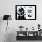 A framed black-and-white poster hanging on a light gray wall above a white and dark wood console shelf, next to a black geometric floor lamp. The poster features a portrait of a person wearing a cap and includes bold text and the number 12.