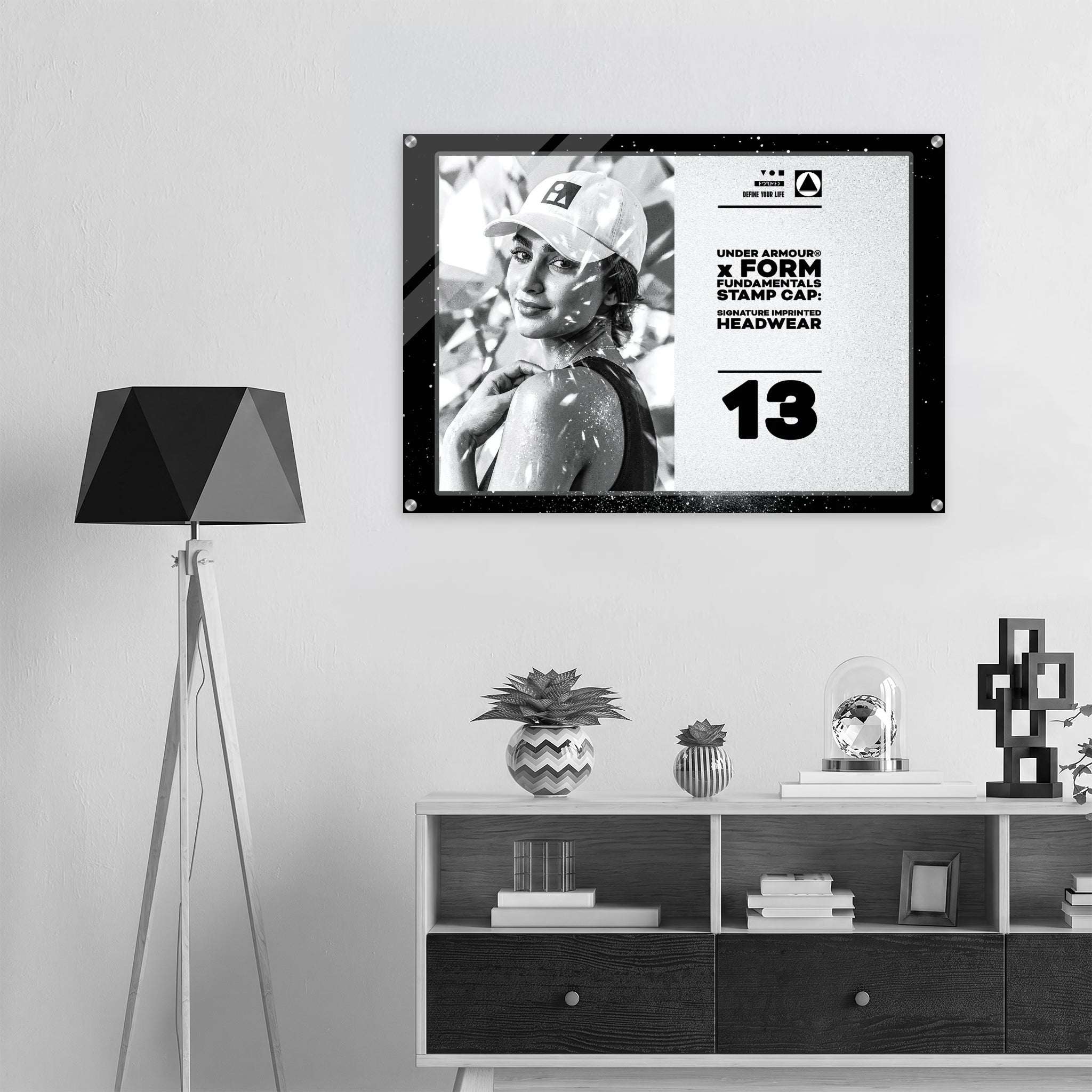 A framed black-and-white poster hangs on a light gray wall above a white and dark wood console shelf, next to a black geometric floor lamp. The poster features a monochrome portrait, bold text from Form Finding Design (FormFD).