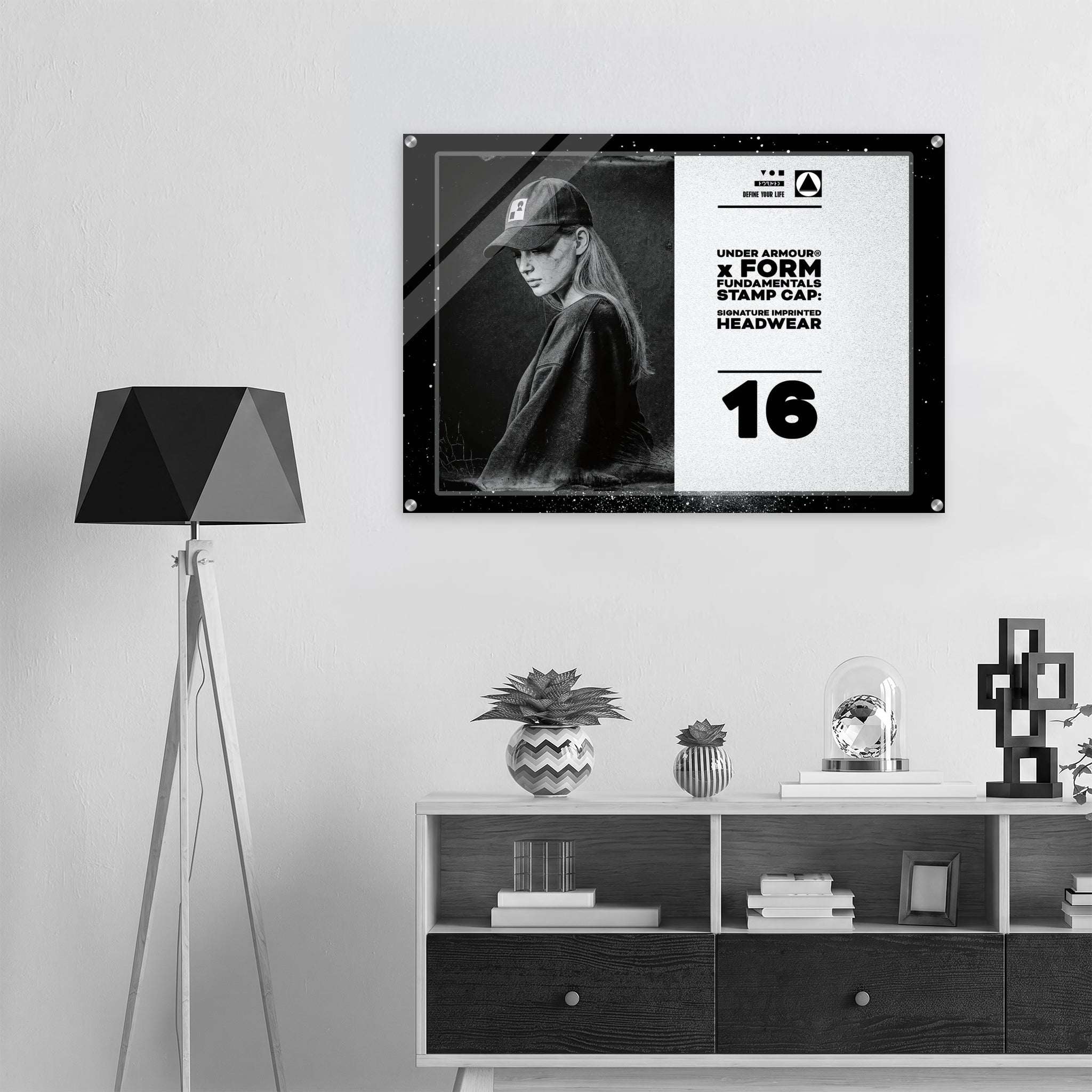 A framed black-and-white poster hanging on a light gray wall above a white and dark wood shelf, next to a black geometric floor lamp. The poster features a woman wearing a cap and bold text from Form Finding Design (FormFD).