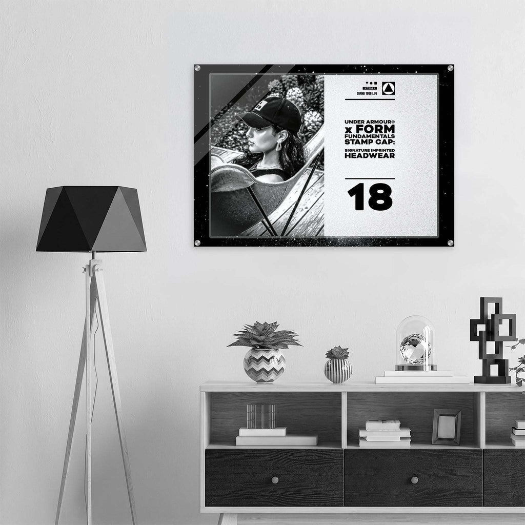 A framed black and white poster hanging on a light gray wall above a white and dark wood shelf, next to a black tripod floor lamp. The poster features a black-and-white photo of a person wearing a cap and the text from Form Finding Design (FormFD).