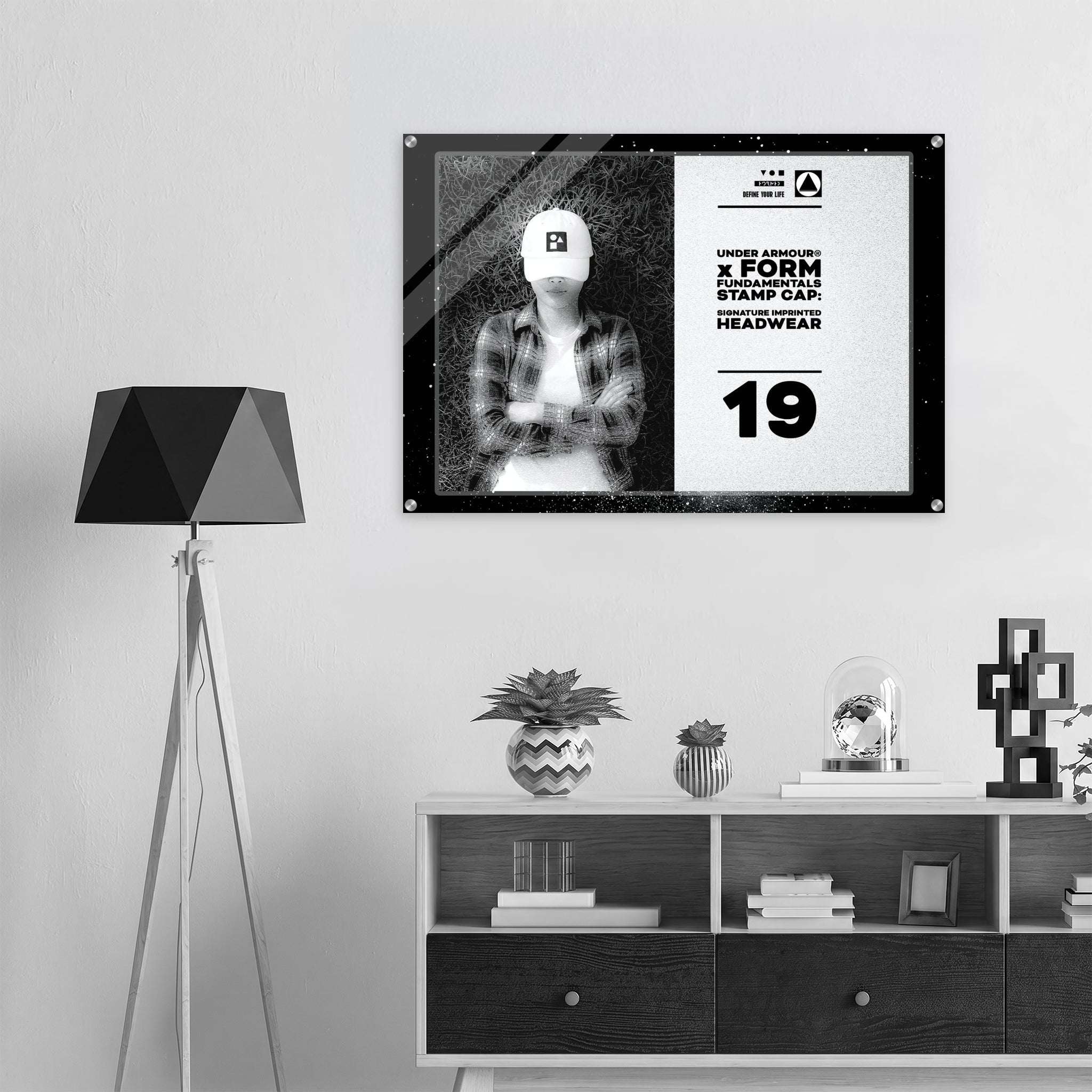 A framed black-and-white poster or print hangs on a light gray wall above a white shelf with decorative objects. The poster shows a split design: a black-and-white photo of a person wearing a cap on the left, and bold text from Form Finding Design (FormFD).