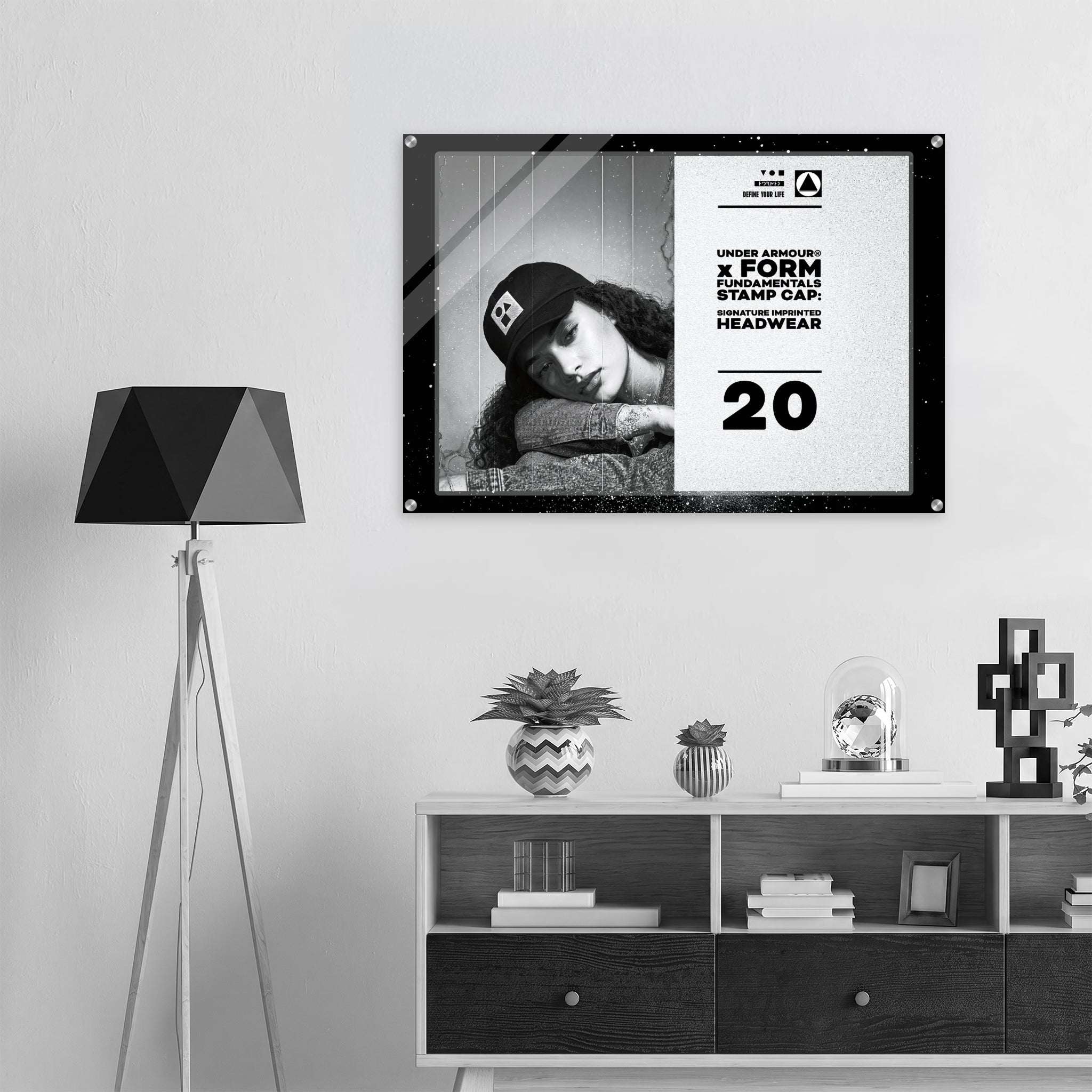 A framed black-and-white poster hanging on a light gray wall above a white and dark wood shelf, next to a black geometric floor lamp. The poster features a woman wearing a cap and bold text from Form Finding Design (FormFD).