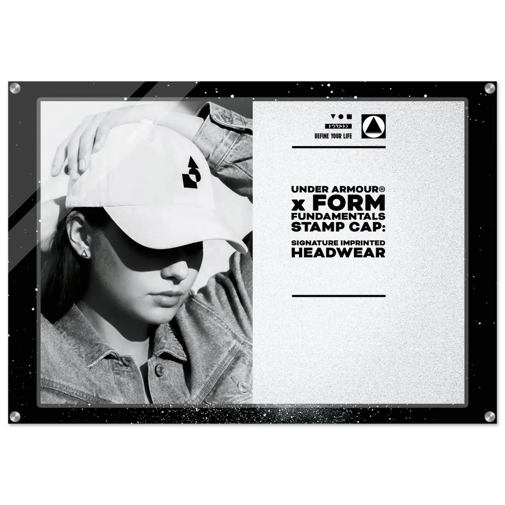 A black-framed promotional display sign featuring a black-and-white photo of a person wearing a white Under Armour cap on the left, and printed text on the right that reads 'UNDER ARMOUR x FORM FUNDAMENTALS STAMP CAP: SIGNATURE IMPRINTED HEADWEAR' with the Under Armour logo. The sign is photographed against a plain white background.