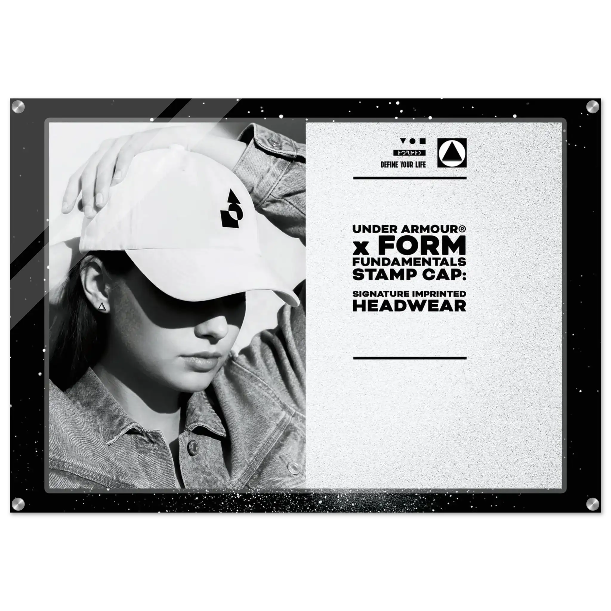 A black-framed promotional display sign featuring a black-and-white photo of a person wearing a white Under Armour cap on the left, and printed text on the right that reads 'UNDER ARMOUR x FORM FUNDAMENTALS STAMP CAP: SIGNATURE IMPRINTED HEADWEAR' with the Under Armour logo. The sign is photographed against a plain white background.