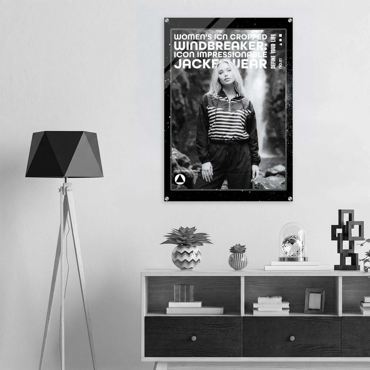A black and white poster featuring a model hanging on a light gray wall in a modern, minimalist room setting with a tripod floor lamp and a wooden shelf below.