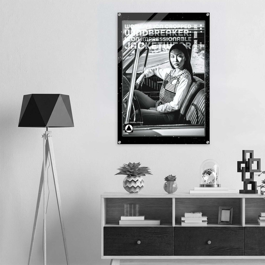 A black and white framed poster featuring a woman in a car with bold text, displayed on a light gray wall above a modern shelf with decorative items and a tripod floor lamp.