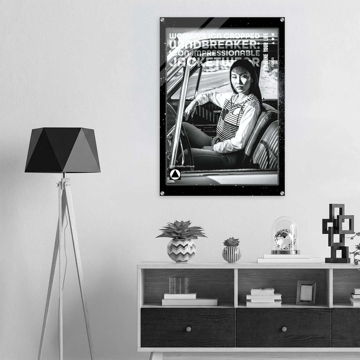 A black and white framed poster featuring a woman in a car with bold text, displayed on a light gray wall above a modern shelf with decorative items and a tripod floor lamp.
