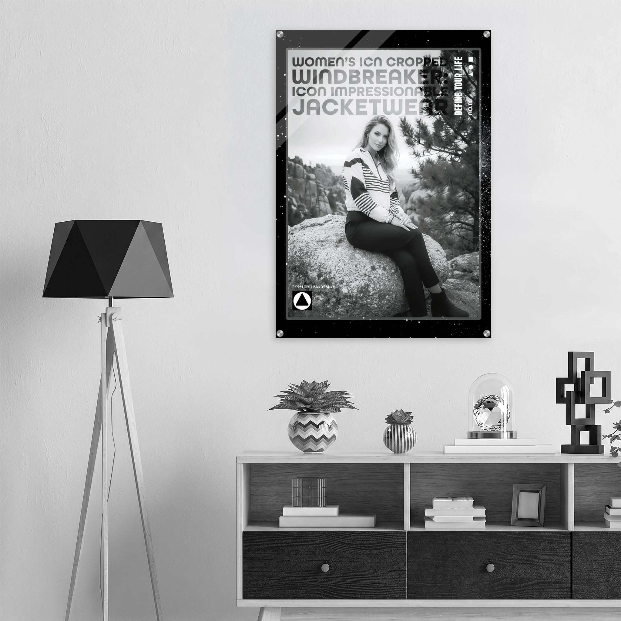 A black and white fashion poster featuring a woman sitting on a rock, with the text 'women's icon cropped windbreaker icon impressionable jacketwear' and a small circular logo, displayed on a wall above a shelf in a modern styled room.