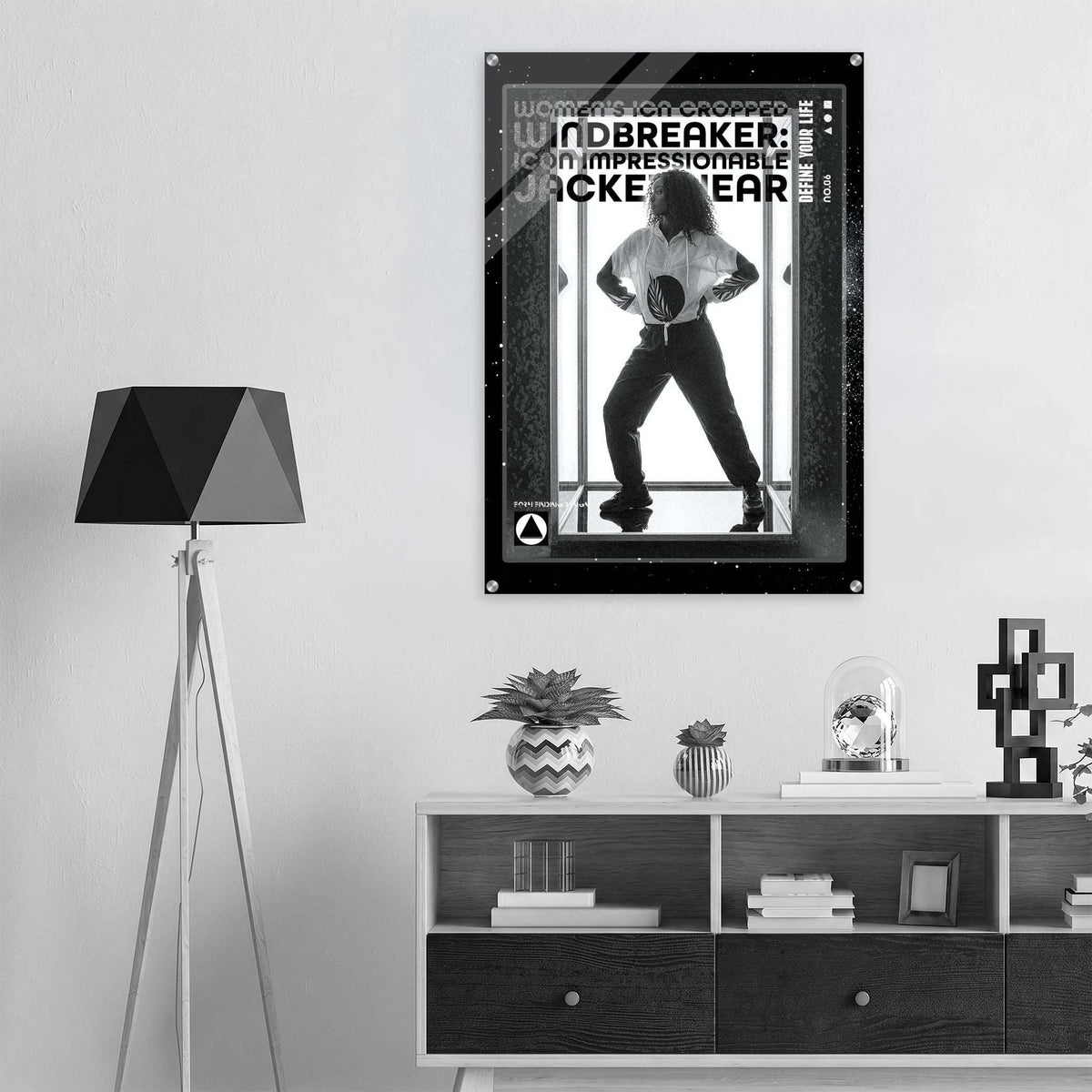 A black and white poster print of 'Windbreaker' jacket hanging on a white wall in a modern, minimalist room setting with a tripod floor lamp and a wooden shelf below.
