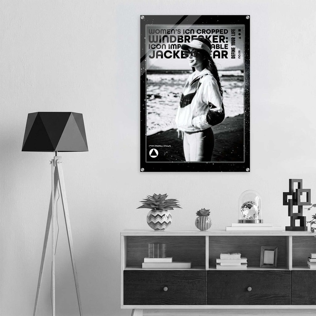 A black and white poster featuring a woman wearing a cap and jacket, with bold text including 'women's icon CROPPED WINDBREAKER: ICON IMPRESSIONABLE JACKETWEAR', displayed on a light gray wall above a shelf in a modern interior setting.