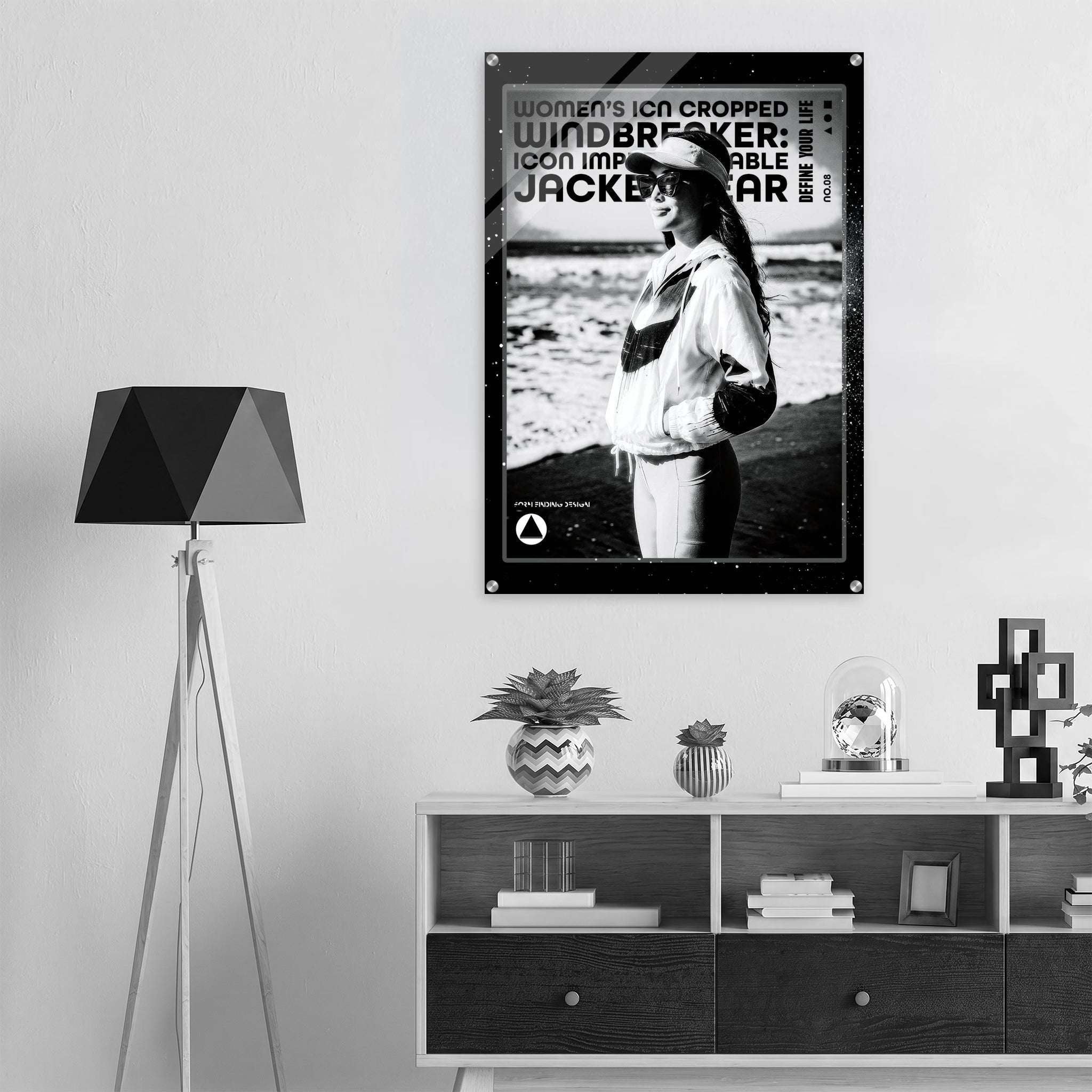 A black and white poster featuring a woman wearing a cap and jacket, with bold text including 'women's icon CROPPED WINDBREAKER: ICON IMPRESSIONABLE JACKETWEAR', displayed on a light gray wall above a shelf in a modern interior setting.