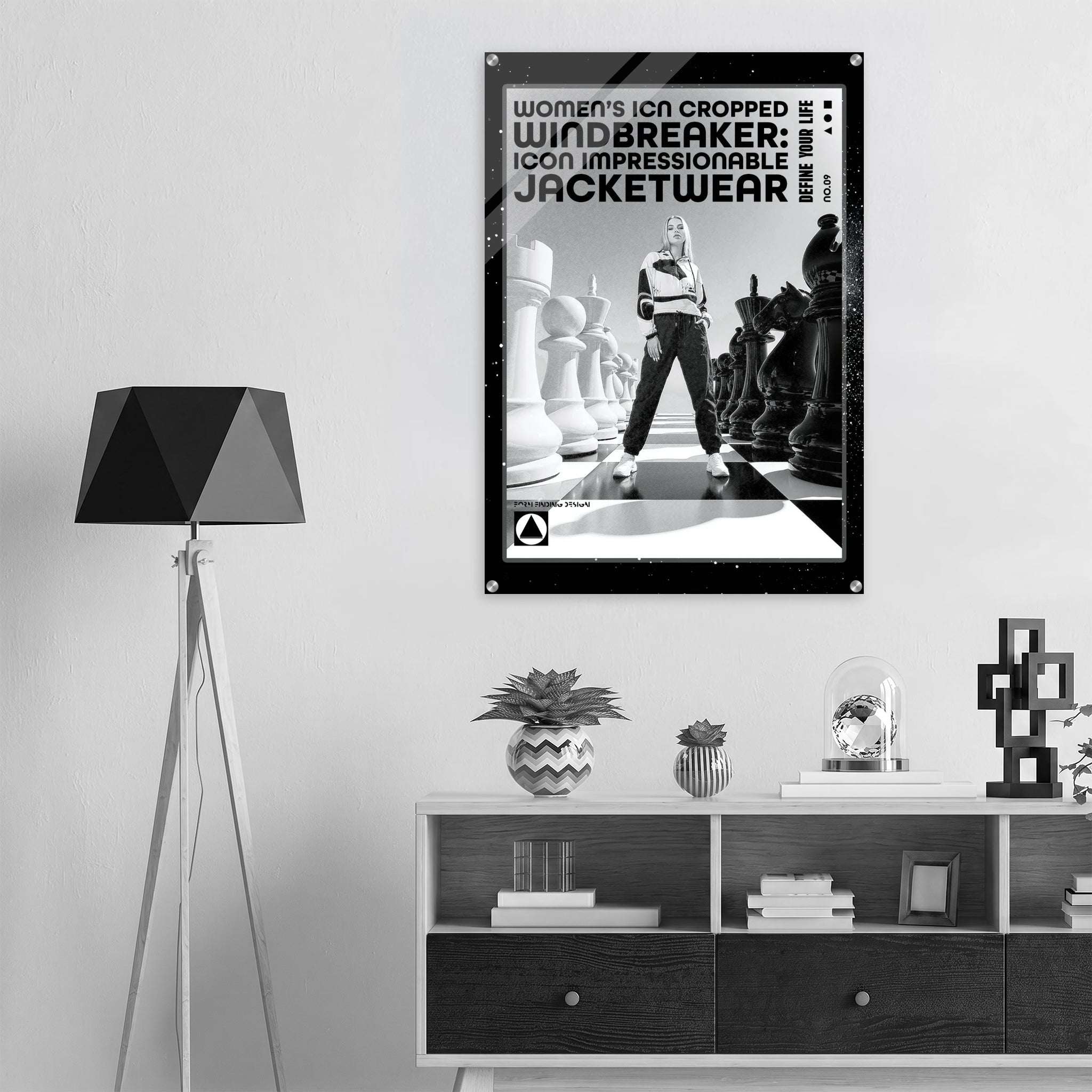 A black and white fashion poster for 'WINDBREAKER: ICON IMPRESSIONABLE JACKETWEAR' hanging on a light gray wall above a wooden shelf in a modern room setting with a tripod floor lamp.