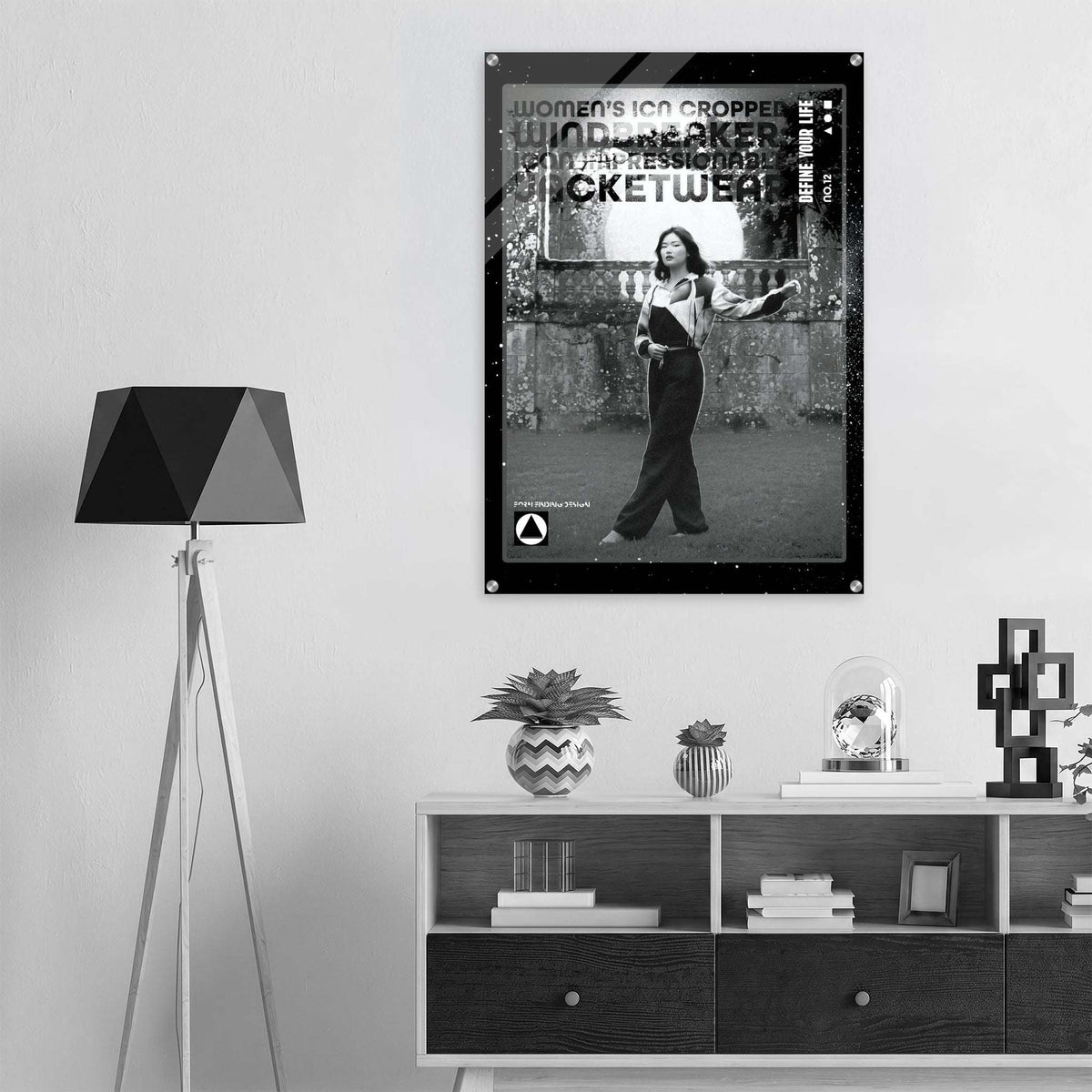 A black and white framed poster of the women's icon cropped windbreaker no.12 displayed on a light gray wall in a modern, minimalist room setting with a tripod floor lamp and a wooden shelf.
