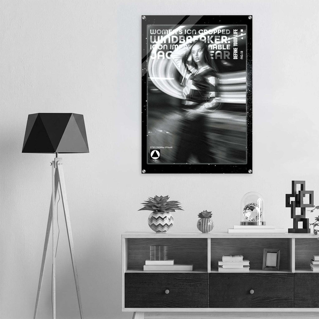 A black and white poster featuring a woman in a motion-blur style graphic with the text 'WOMEN'S ICON CROPPED WINDBREAKER' is displayed on a light gray wall above a modern shelf with decorative items and a tripod floor lamp.