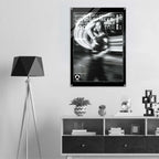 A black and white poster featuring a woman in a motion-blur style graphic with the text 'WOMEN'S ICON CROPPED WINDBREAKER' is displayed on a light gray wall above a modern shelf with decorative items and a tripod floor lamp.