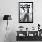 A black and white framed poster hangs on a light gray wall above a modern white and dark wood console table. The room is styled with a black tripod floor lamp on the left, decorative plants, books, and a glass dome.