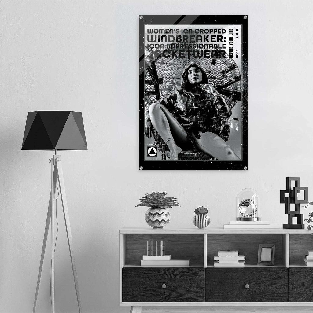 A black and white framed poster hanging on a light gray wall above a wooden shelf, showing a seated woman in athletic wear with bold text reading 'Women's Icon Cropped Windbreaker: Impressionable Jacketwear.' The room includes a black floor lamp on the left and decorative objects on the shelf below.