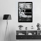 A black and white poster featuring a screen printed and burned model hanging on a light gray wall in a modern, minimalist room setting with a tripod floor lamp and a low wooden shelf.