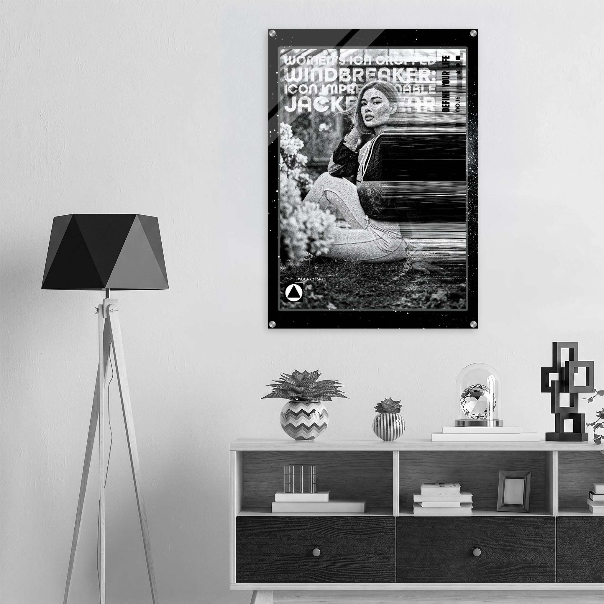 A black and white poster featuring a screen printed and burned model hanging on a light gray wall in a modern, minimalist room setting with a tripod floor lamp and a low wooden shelf.