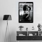 A black and white framed poster featuring a woman and bold text hangs on a light gray wall above a white shelf with decorative objects. A black tripod floor lamp stands to the left.