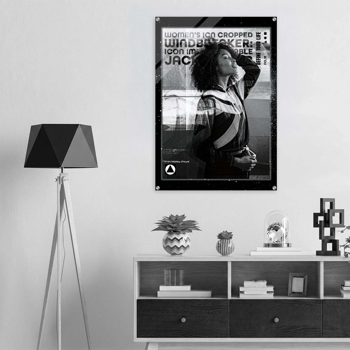 A black and white framed poster featuring a woman and bold text hangs on a light gray wall above a white shelf with decorative objects. A black tripod floor lamp stands to the left.