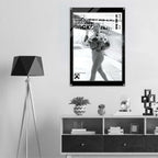 A black and white poster featuring a woman in a street scene with bold text, displayed on a light gray wall above a modern wooden shelf with decorative items. A black tripod floor lamp stands to the left.
