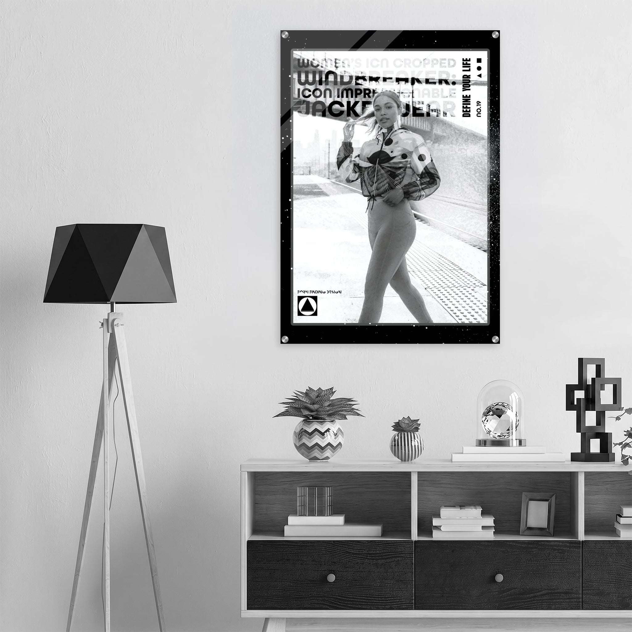 A black and white poster featuring a woman in a street scene with bold text, displayed on a light gray wall above a modern wooden shelf with decorative items. A black tripod floor lamp stands to the left.