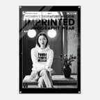 A black and white fashion poster featuring a seated woman in a hoodie with bold text reading 'WOMEN'S SIGNATURE HOODIE: IMPRINTED ICONOGRAPHY WEAR.' The poster is shown hanging against a light gray wall, with visible metal corner hardware.