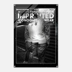 A black and white fashion poster print featuring a stylized figure in front of a corrugated metal wall, with bold text reading 'WOMEN'S SIGNATURE HOODIE' and 'IMPRINTED ICONOGRAPHY WEAR'. The poster is shown hanging against a light gray wall.