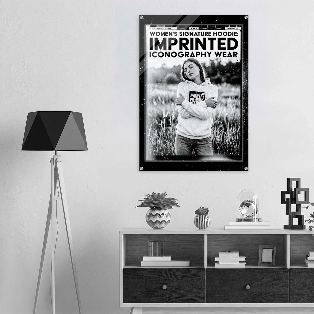 A black and white poster print featuring a woman in a hoodie in a field, with bold text reading 'WOMEN'S SIGNATURE HOODIE IMPRINTED ICONOGRAPHY WEAR', displayed on a wall above a modern console table in a minimalist room setting.