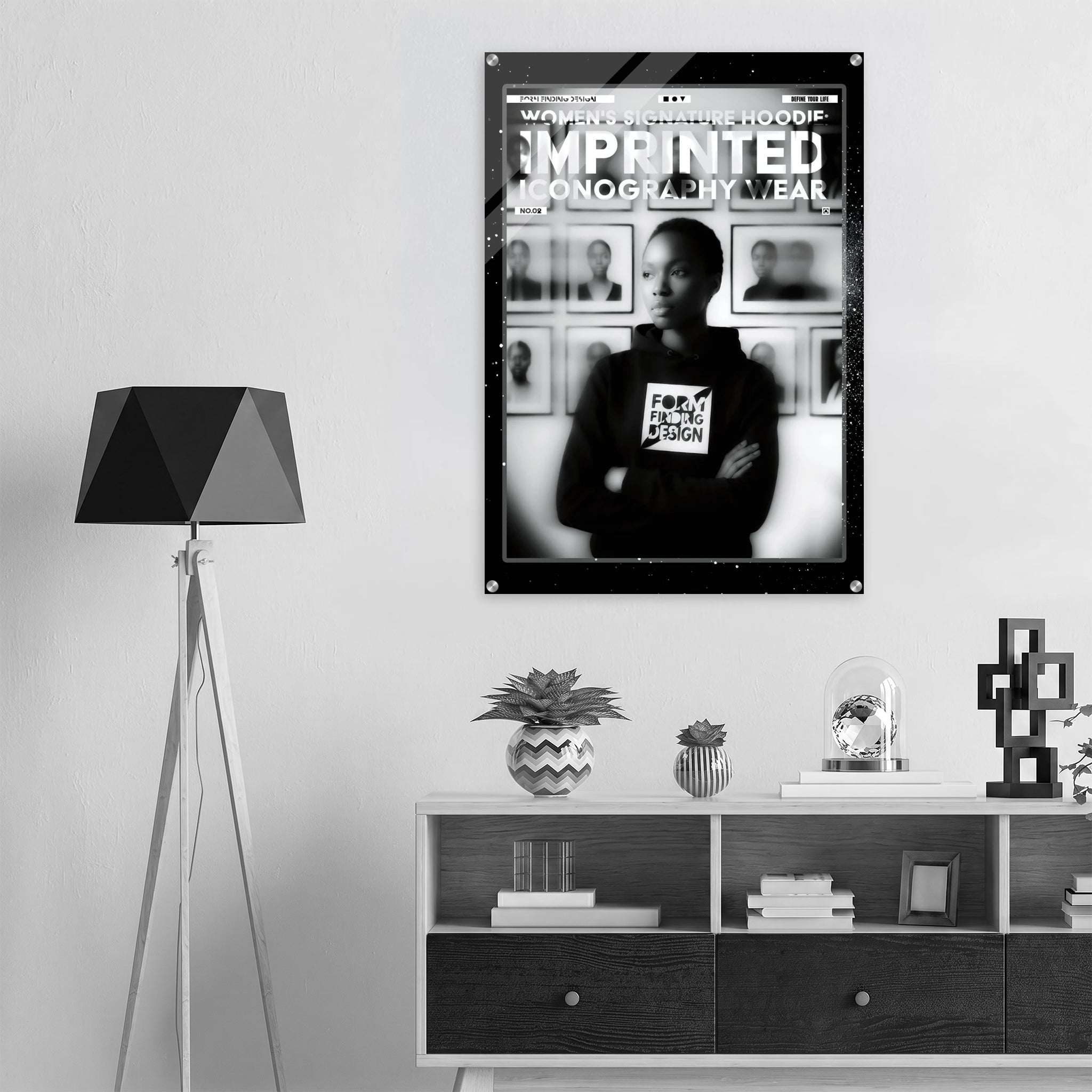 A black and white framed poster hanging on a light gray wall above a white shelf with books and decor. The poster features a person wearing a hoodie with the text 'FORM FINDING DESIGN' and the headline 'WOMEN'S SIGNATURE HOODIE IMPRINTED ICONOGRAPHY WEAR'.