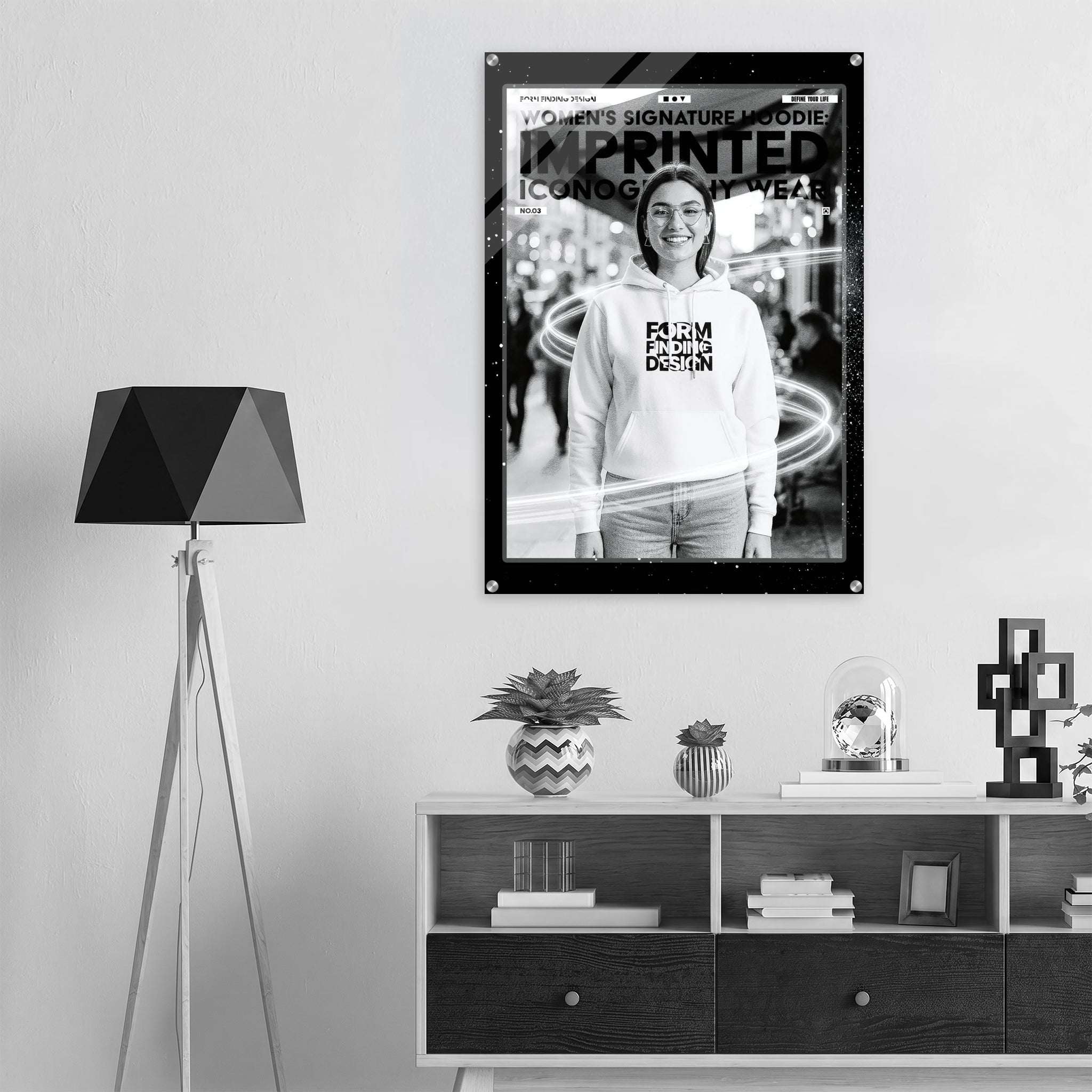 A black and white poster print featuring a woman wearing a hoodie with text, displayed on a light gray wall above a modern shelf with decorative objects and a tripod floor lamp.