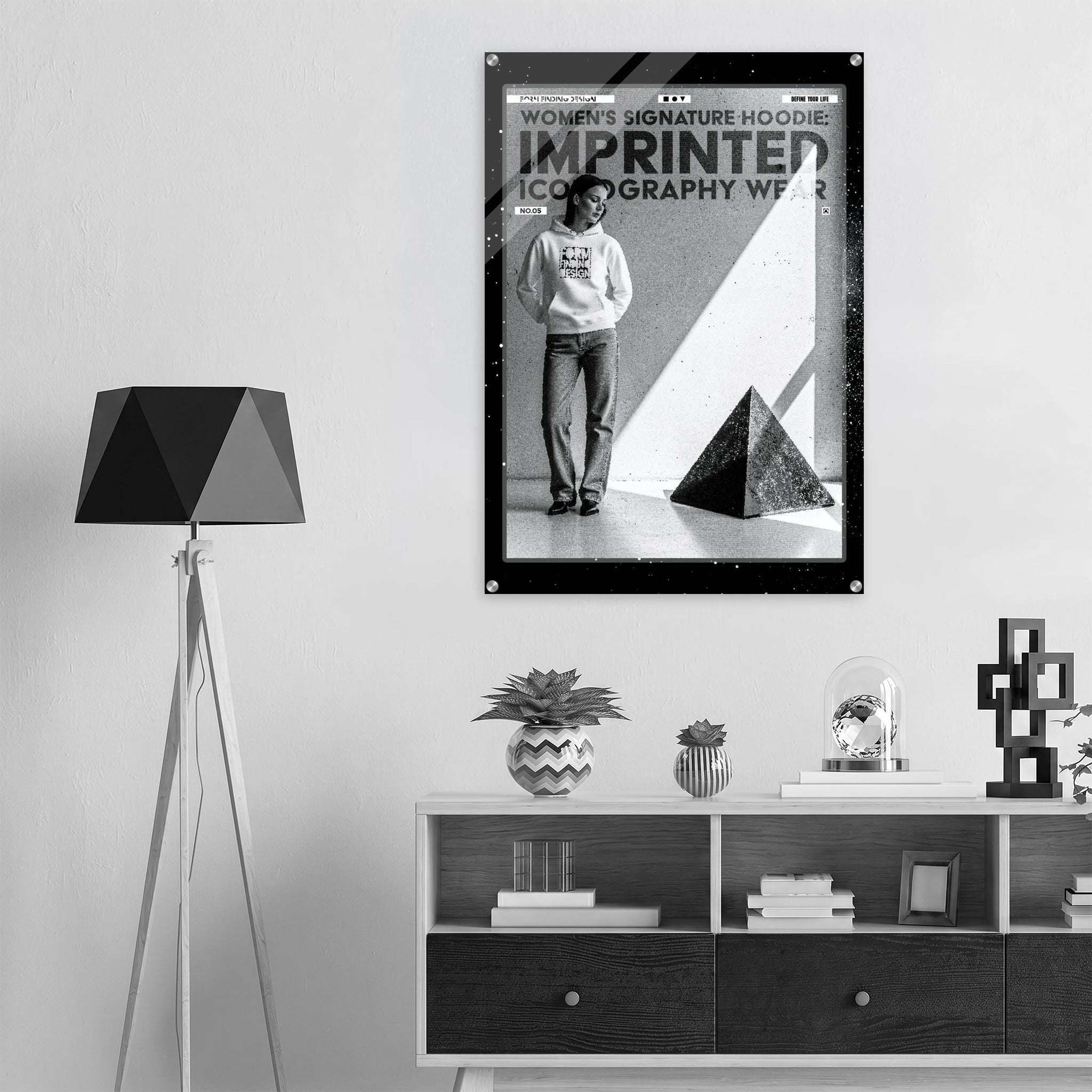 A black-framed wall art print hangs above a white cabinet in a modern room. The print features a black-and-white fashion poster with the text 'WOMEN'S SIGNATURE HOODIE IMPRINTED ICONOGRAPHY WEAR' and a photo of a person wearing a hoodie.