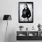 A black and white fashion poster hanging on a light gray wall above a wooden shelf, featuring a woman in a black hoodie with the text 'WOMEN'S SIGNATURE HOODIE IMPRINTED ICONOGRAPHY WEAR'. The room includes a tripod floor lamp on the left and decorative objects on the shelf.