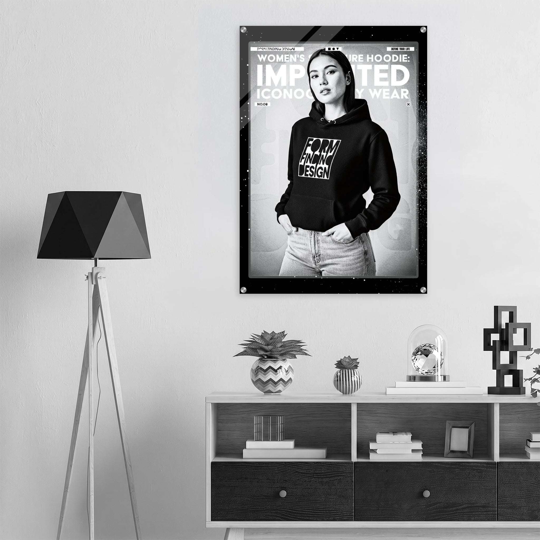 A black and white fashion poster hanging on a light gray wall above a wooden shelf, featuring a woman in a black hoodie with the text 'WOMEN'S SIGNATURE HOODIE IMPRINTED ICONOGRAPHY WEAR'. The room includes a tripod floor lamp on the left and decorative objects on the shelf.