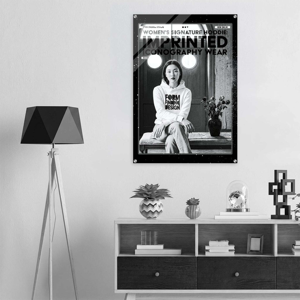 A black and white fashion poster for 'Women's Signature Hoodie Imprinted Iconography Wear' hangs on a light gray wall above a modern white and dark wood sideboard. The room is styled with a black geometric tripod floor lamp on the left, decorative plants, books, and a small glass dome on the cabinet.