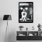 A black and white fashion poster for 'Women's Signature Hoodie Imprinted Iconography Wear' hangs on a light gray wall above a modern white and dark wood sideboard. The room is styled with a black geometric tripod floor lamp on the left, decorative plants, books, and a small glass dome on the cabinet.