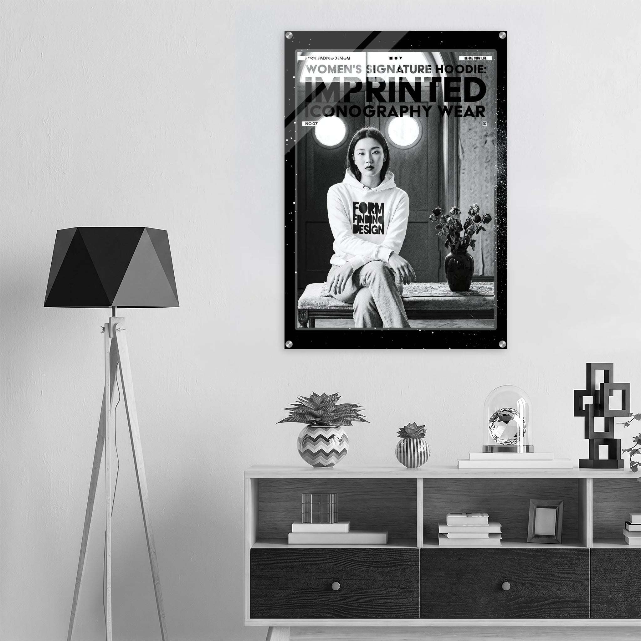 A black and white fashion poster for 'Women's Signature Hoodie Imprinted Iconography Wear' hangs on a light gray wall above a modern white and dark wood sideboard. The room is styled with a black geometric tripod floor lamp on the left, decorative plants, books, and a small glass dome on the cabinet.