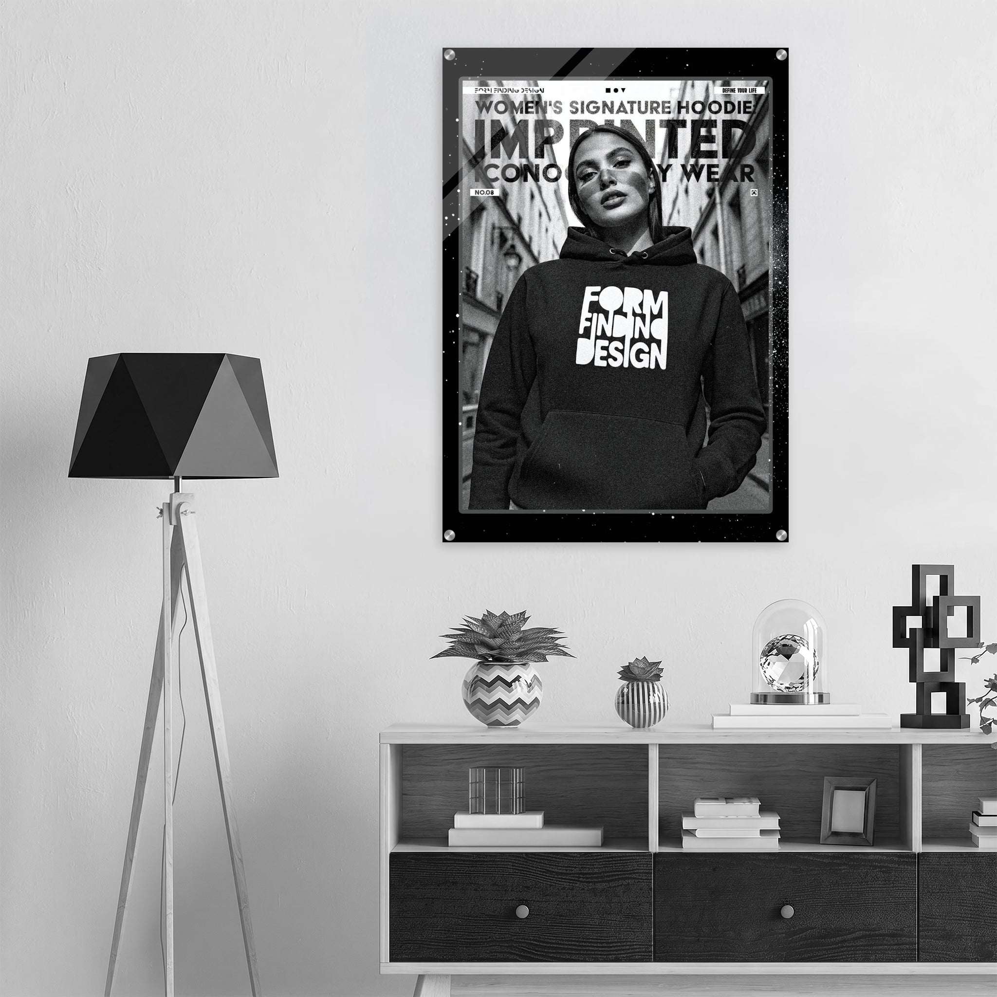 A black and white fashion poster hanging on a light gray wall above a white and dark wood console shelf. The poster features a woman wearing a hoodie with the text 'FORM FINDING DESIGN' and 'WOMEN'S SIGNATURE HOODIE'.